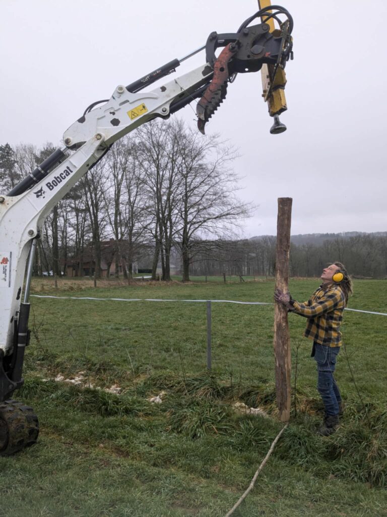Un homme en habit de travail tient un poteau en bois pendant qu'une machine plante le poteau en bois dans le sol