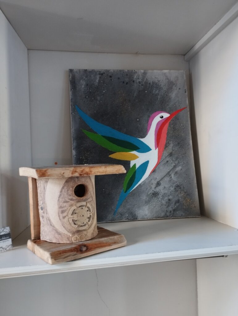 Un nichoir en terre cuite avec des motifs abstraits dessinés dessus en avant plan. En arrière plan, la peinture d'un colibri.