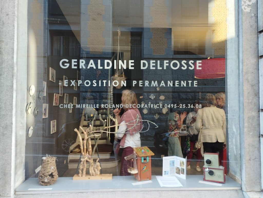 La vritrine d'une salle d'exposition avec l'inscription "Géraldine Delfosse: Exposition permanente". On peut apperçevoir à travers la vitre des personnes dans la salle ainsi que des oeuvres exposées dans l'étalage.