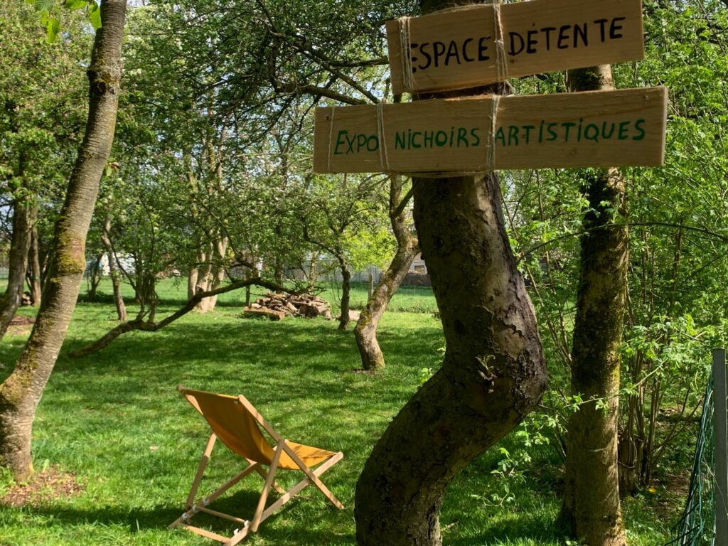 En arrière plan un transat dans un verger
En avant plan, deux panneaux en bois accrochés sur un tronc d'arbre.
Sur le premier est inscrit "Espace détente" et sur le second "Expo nichoirs artistiques"