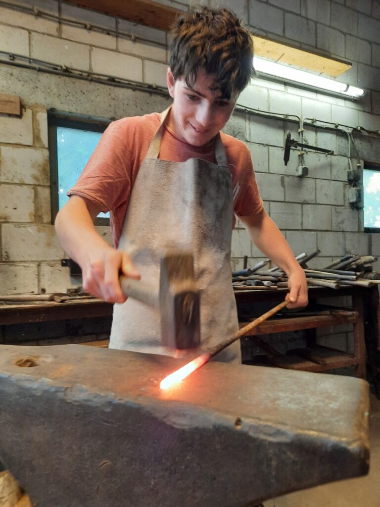 un adolescent forge un outil avec un marteau et une enclume