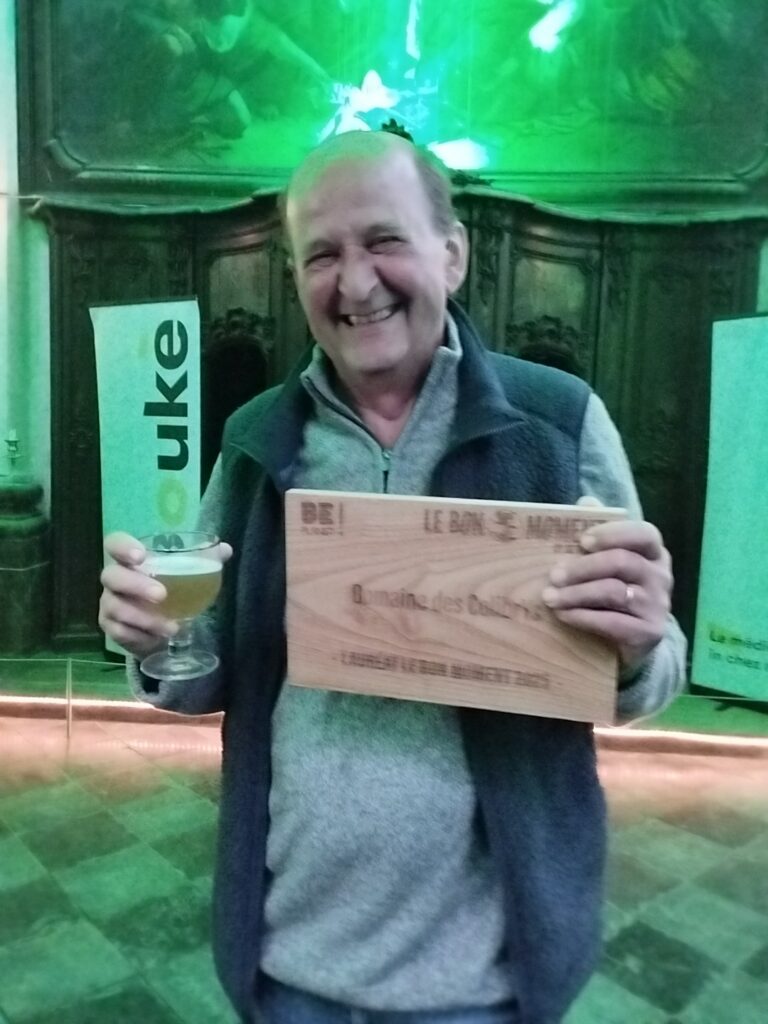 Un homme d'une soixantaine d'années tient une bière dans sa main droite et une pancarte en bois avec "Le Domaine de ColibrYs" gravé dessus