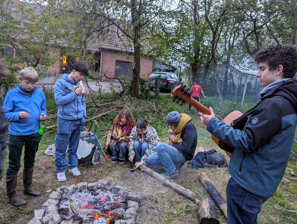 Groupe d'adolecsents qui mange autour d'un feu de camp. L'un d'eux joue de la guitare